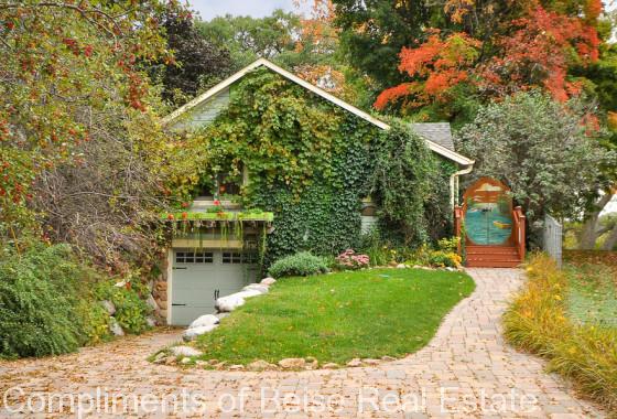 A house at 5005 Avon Drive features a garage partially covered in ivy, a brick driveway, and a painted mural on the side entrance, all surrounded by trees with vibrant autumn foliage. - Beise Real Estate