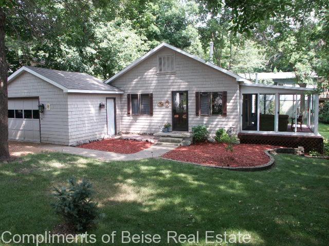 Single-story house at 1736 Sumach Lane with attached garage, gray siding, and large windows, surrounded by trees and a lawn with red mulch landscaping. - Beise Real Estate