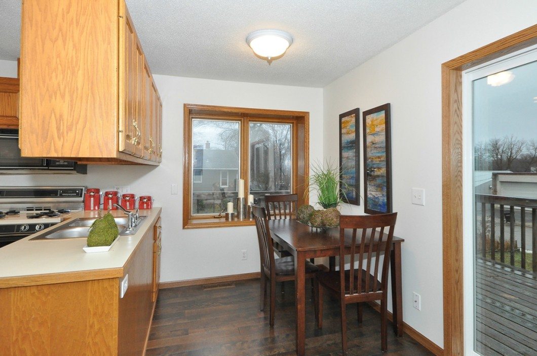 A small dining area with a wooden table and four chairs sits by the window, adjacent to a kitchen with wooden cabinets and stainless steel appliances in this cozy Fairview Lane home in Mound. - Beise Real Estate