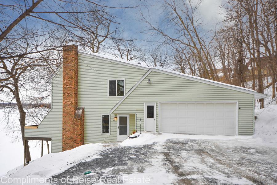 Charming two-story house at 3035 Bluffs Lane with light green siding, brick chimney, and attached two-car garage, nestled on a snowy hillside amid bare winter trees. - Beise Real Estate