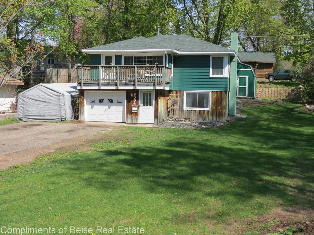 A two-story green house at 5004 Jennings Rd features a wooden balcony, garage, and a large front yard with grass and trees in the background. - Beise Real Estate