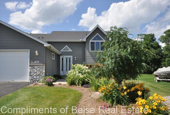 Single-story gray house with stone accents on Hidden Creek Blvd, featuring a front garden with yellow flowers and shrubs, a manicured lawn, and a blue sky with scattered clouds. - Beise Real Estate