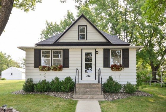 Charming small white house with black shutters at 312 Bluejay Ave, featuring central front steps, flower boxes under windows, and neatly trimmed bushes, all set on a grassy lawn with trees in the background. - Beise Real Estate