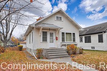 A small, single-story house with white stucco exterior on 3409 44th Ave South features a front porch and concrete steps, surrounded by autumn leaves and bare trees under a partly cloudy sky. - Beise Real Estate
