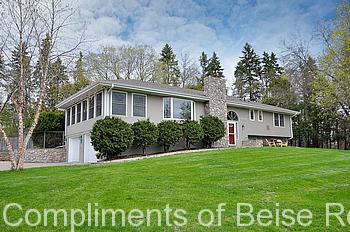 Charming single-story house at 2020 W 65th Street with stone chimney, gray siding, red door, and large windows, surrounded by a green lawn and trees under a partly cloudy sky. - Beise Real Estate