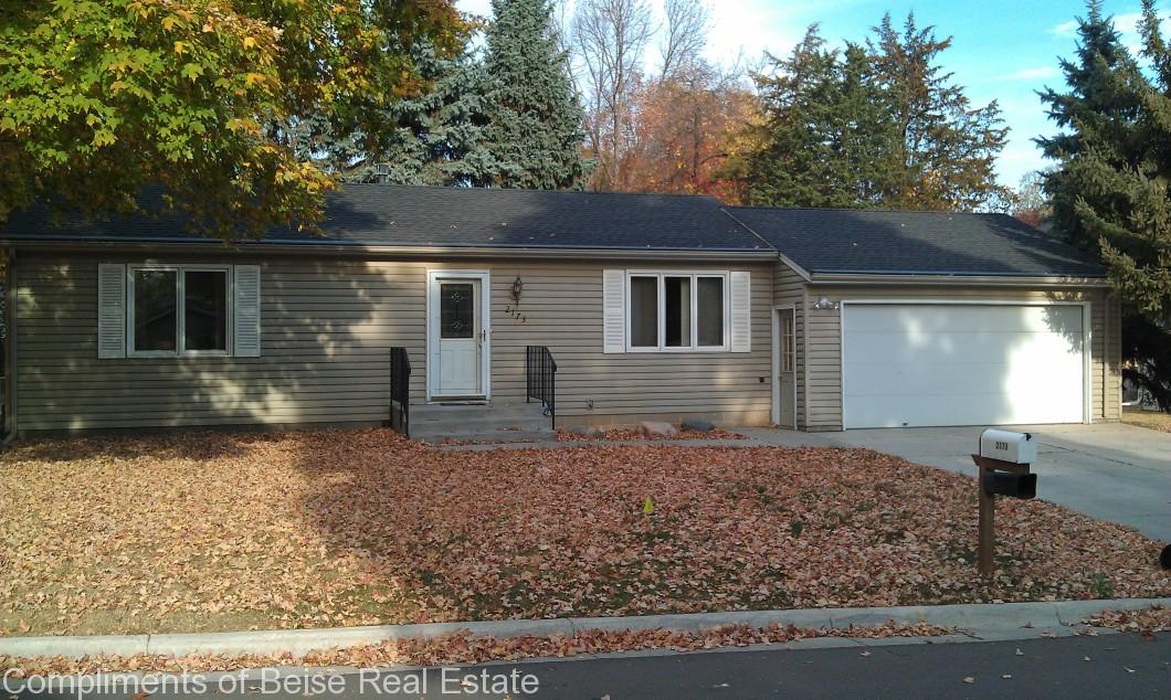 Charming single-story real estate on Cardinal Lane with beige siding, double garage, and a small porch, surrounded by autumn leaves covering the lawn and driveway. - Beise Real Estate