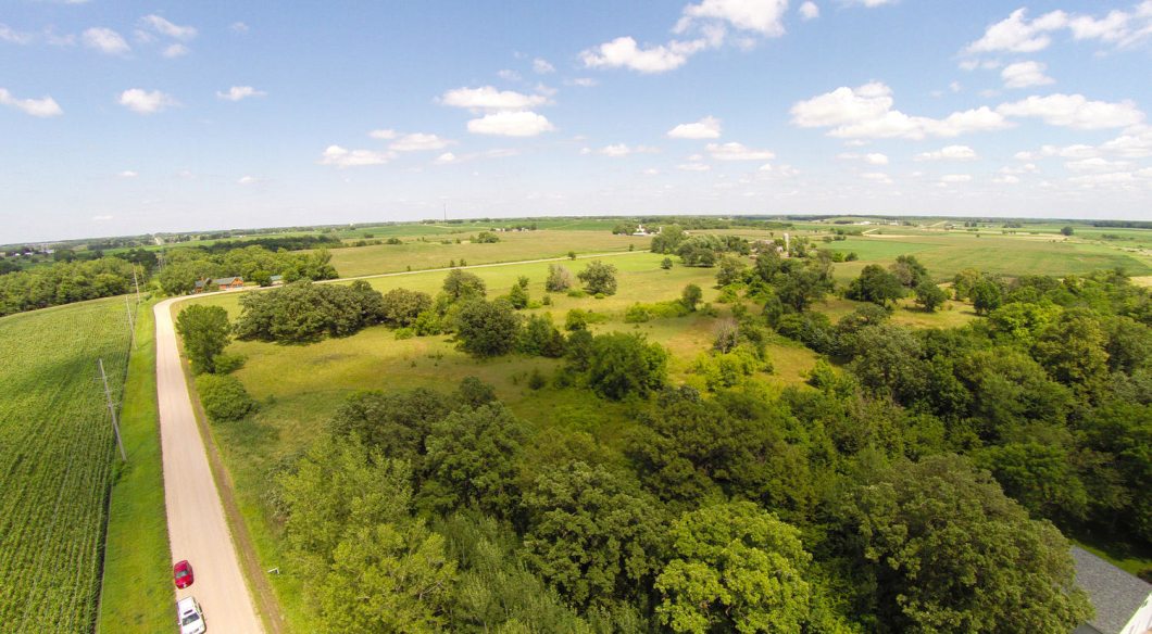 A rural landscape near Howard Lake with green fields, scattered trees, and a dirt road running alongside farmland under a partly cloudy sky. - Beise Real Estate