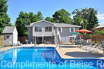 A backyard pool area at 21132 Butternut St. NW features patio furniture, a large umbrella, and a two-story gray house with a deck in the background. Trees and a small shed are visible near the fence. - Beise Real Estate