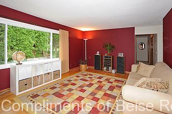 A living room at 2245 Aquila Ave N features red walls, a large window, beige couch, patterned area rug, white storage unit, and lush houseplants. - Beise Real Estate