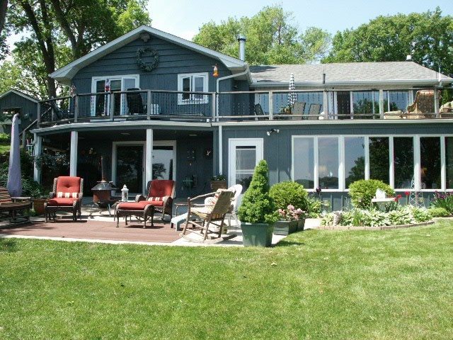 Two-story gray house at 1786 Shorewood Lane with large windows, an upper balcony with patio furniture, and a landscaped garden in front of a grassy lawn. - Beise Real Estate