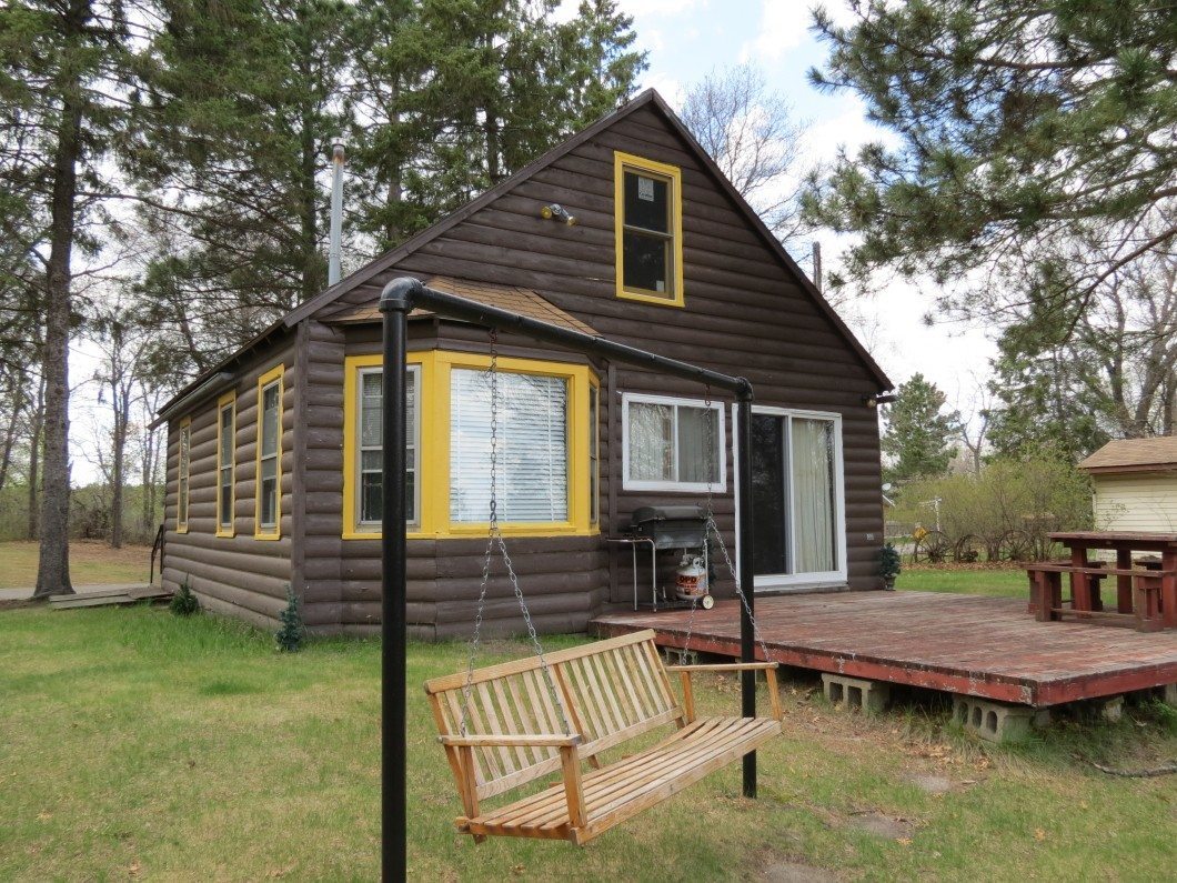A cozy brown Cabin with yellow trim in Big Lake Township, featuring a wooden swing, a deck with a picnic table, and surrounded by trees. - Beise Real Estate