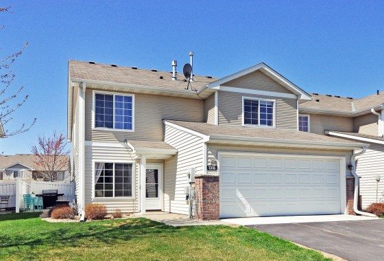 Two-story suburban home at 1318 Pembrooke Lane with beige siding, brick accents, and a double garage under a clear blue sky. - Beise Real Estate