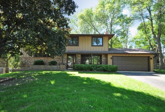 Two-story suburban house at 9009 44th Circle N with a brown exterior, large windows, and attached two-car garage, surrounded by green lawn and mature trees. - Beise Real Estate
