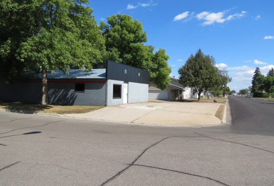 A gray, single-story commercial building with a flat roof sits on a corner lot at 211 Central Avenue, featuring a concrete driveway and nearby trees under a partly cloudy sky. - Beise Real Estate