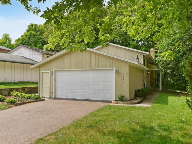 Single-story house at 3149 Alexander Lane featuring an attached two-car garage, beige siding, and a driveway, all surrounded by a lush green lawn and mature trees. - Beise Real Estate