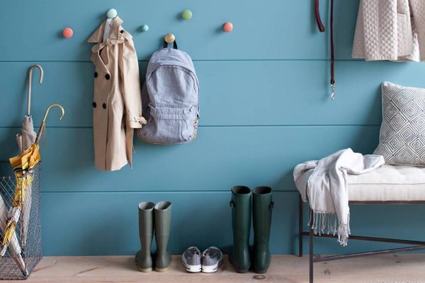 A blue wall with colorful hooks holds a beige coat and gray backpack; below are rain boots and sneakers. An umbrella, bench, and hanging clothes complete this organized entryway, making being organized simple every day. - Beise Real Estate