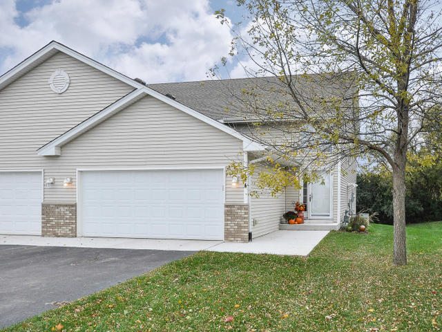 A light-colored suburban duplex at 144 Pheasant Ridge Drive with a two-car garage, autumn leaves on the ground, and pumpkins by the front door—an inviting real estate opportunity. - Beise Real Estate