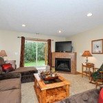 A living room at 144 Pheasant Ridge features brown sofas, a wooden coffee table, a fireplace with a TV above, two lamps, a rocking chair, and sliding glass doors opening to a green yard—perfect for your next real estate move. - Beise Real Estate