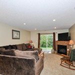 Welcome to 144 Pheasant Ridge Drive, featuring a spacious living room with a large sectional sofa, carpeted floor, table lamps, a cozy fireplace with TV above, rocking chair, and sliding glass doors opening to the outdoors. - Beise Real Estate