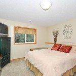 Bedroom at 144 Pheasant Ridge features a bed with two red pillows, a green dresser topped with a TV, neutral walls adorned with decor, and a vase of branches—an inviting real estate opportunity on Pheasant Ridge Drive. - Beise Real Estate