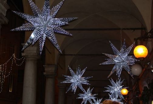 Large illuminated star-shaped decorations hang from the ceiling of an arched walkway, accompanied by round orange lights and strings of small fairy lights—perfect inspiration for your holiday planning. - Beise Real Estate