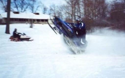 A person performs a wheelie on a snowmobile in a snowy yard, while another rides near a house—maybe start looking for a home where winter fun is right outside your door. - Beise Real Estate