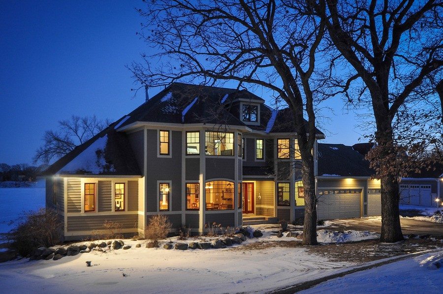 A two-story house with illuminated windows is shown at dusk, surrounded by leafless trees and a snow-covered yard, while warm lighting from the three-car garage on the right helps light up your life. - Beise Real Estate