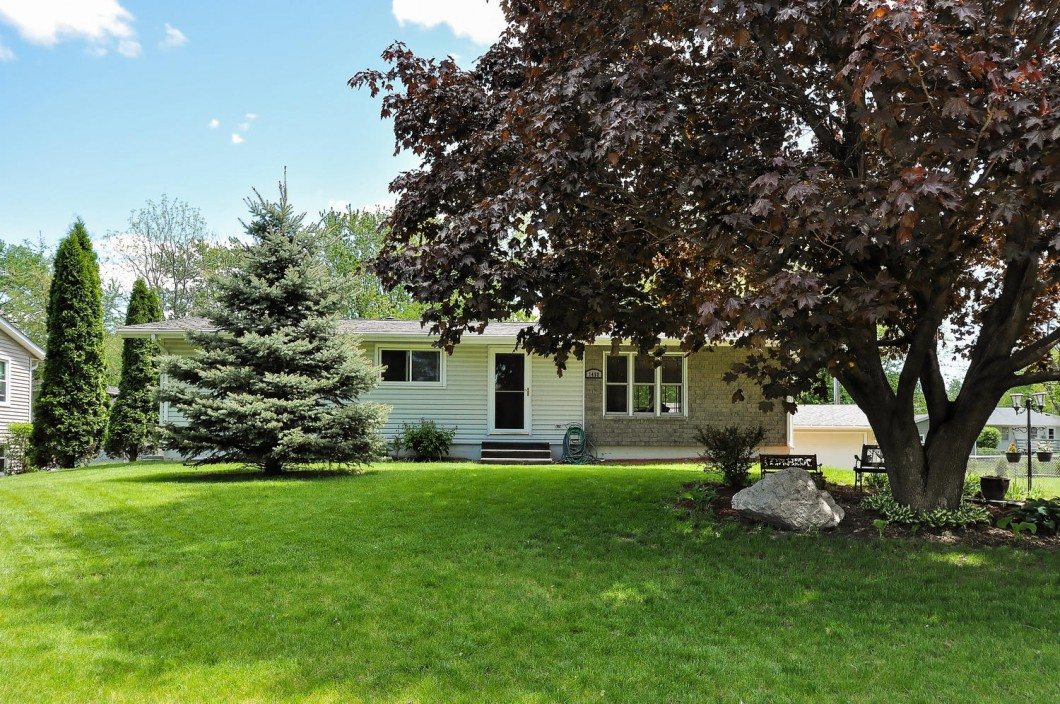 Single-story house with light siding on Rainbow Avenue, a black front door, large windows, and a shaded front yard featuring a mature tree and well-kept lawn in Maple Plain. - Beise Real Estate