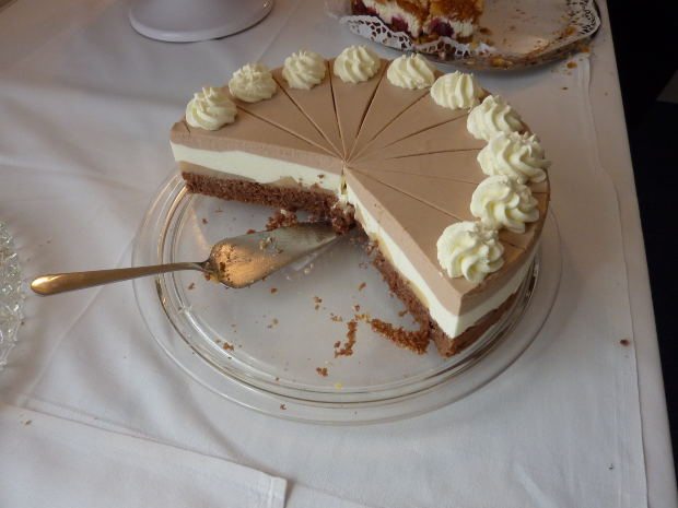 A partially sliced layered cake with swirls of cream on top sits on a glass platter with a serving utensil on a white tablecloth, creating a scene as inviting as good houses in the housing market. - Beise Real Estate