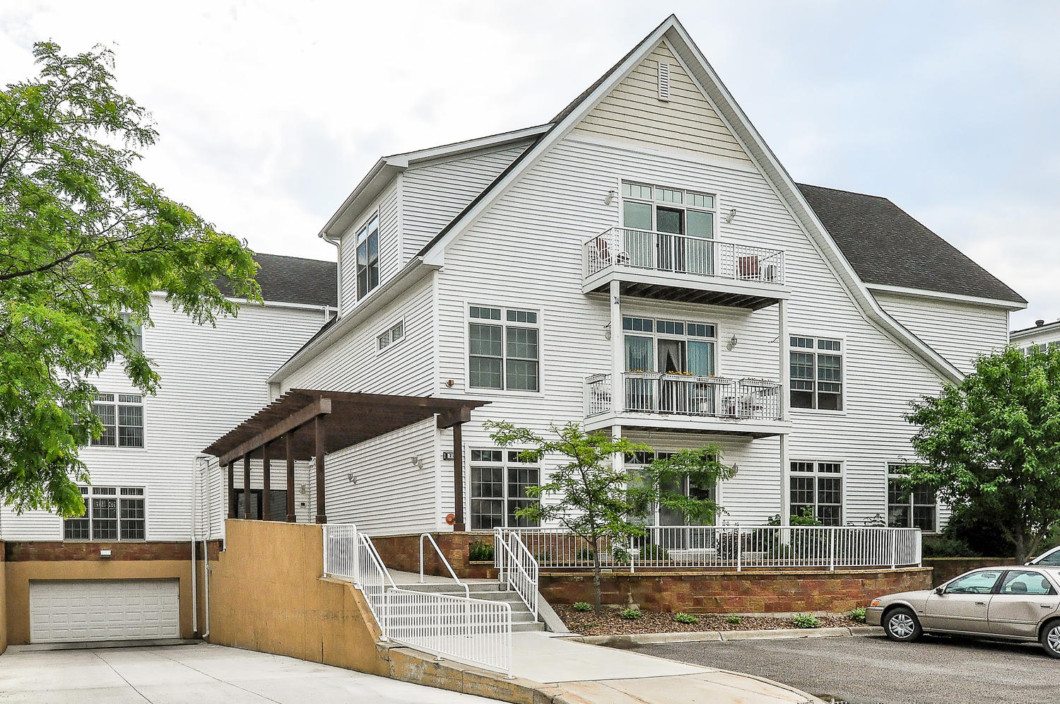 A modern white three-story apartment building at 2100 Old School Rd features balconies, large windows, a wooden pergola, a ramp, and a car parked nearby. - Beise Real Estate