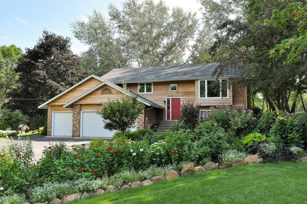 A split-level house at 21525 Treeline Drive features light wood siding, a red front door, and a two-car garage, surrounded by lush landscaping and blooming flowers. - Beise Real Estate