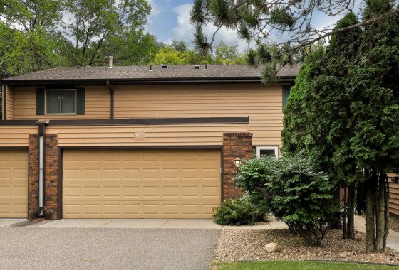 Two-car garage attached to a tan, two-story townhouse with brown trim at 4729 Spring Circle, surrounded by trees, shrubs, and a small gravel area near the driveway. - Beise Real Estate