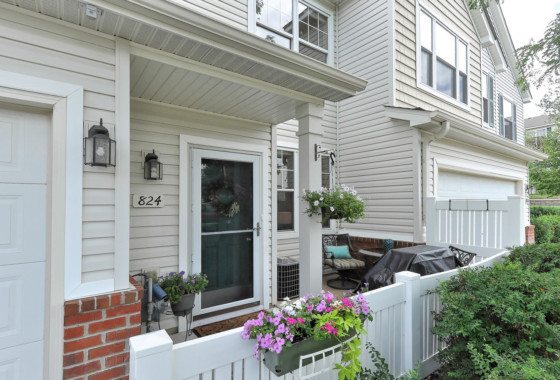 A townhouse front entrance at 824 Princeton Avenue features a glass storm door, potted plants, a hanging basket, white fence, and house number 824. - Beise Real Estate