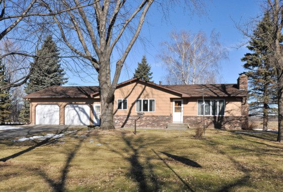 Single-story ranch house at 6478 Desoto Avenue NW with attached two-car garage, tan siding, and brick accents, surrounded by leafless trees and a patchy winter lawn under a clear blue sky. - Beise Real Estate