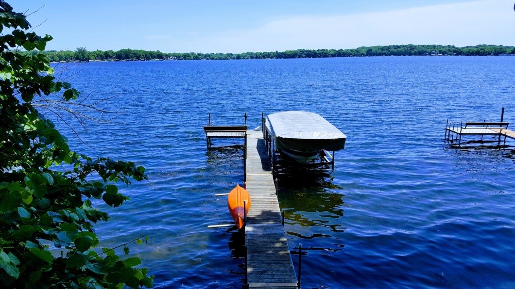 A wooden dock extends into a blue lake at 2960 Highland Court, with a covered boat and an orange kayak beside it. Trees and two more docks are visible in the background. - Beise Real Estate