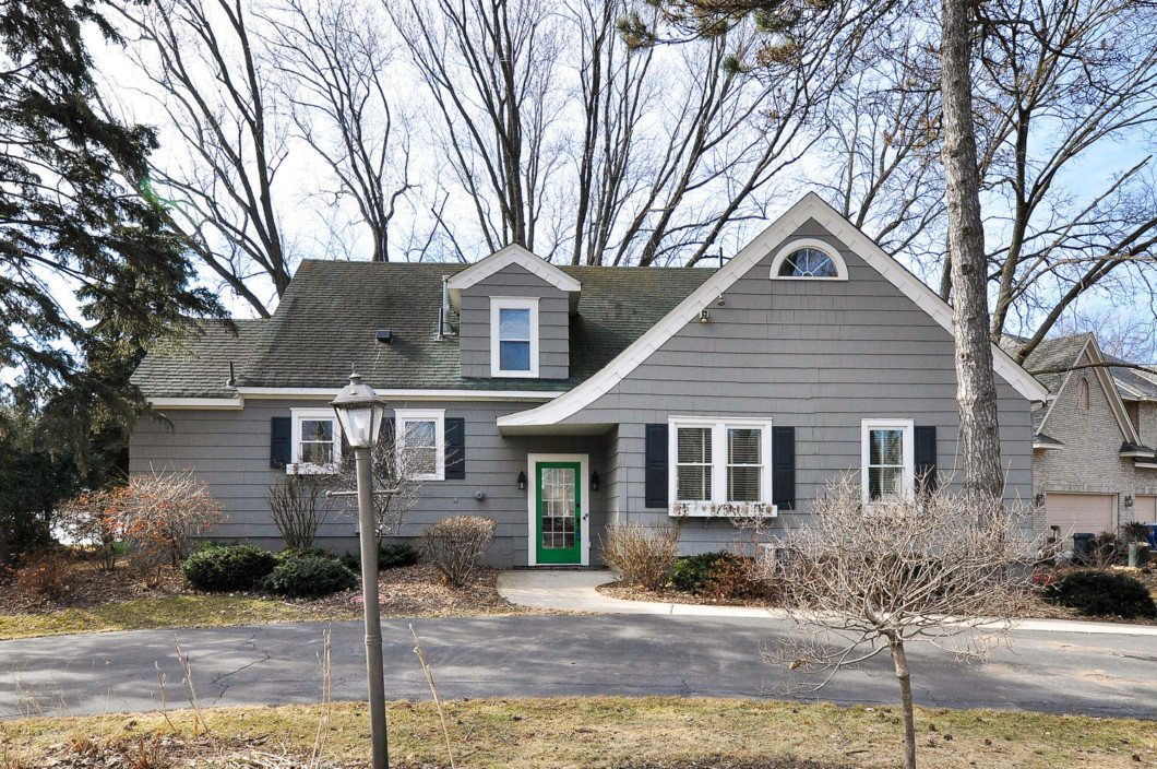 A gray two-story house with white trim, a green front door, dormer windows, and a curved driveway sits at 2625 Wilshire Blvd, surrounded by trees and shrubs. - Beise Real Estate