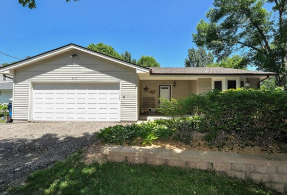Single-story house at 2116 Grandview Court with light siding, a double garage door, front porch, and landscaped yard with shrubs and trees under a clear blue sky. - Beise Real Estate
