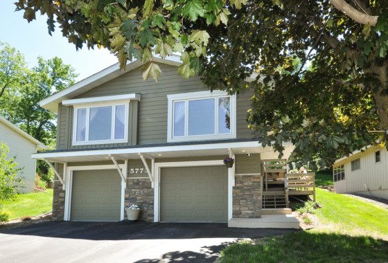 A two-story house with green siding, stone accents, and two garage doors at 5773 Grandview Blvd, partially shaded by a large tree in the foreground. The house number 577 is visible above the garage. - Beise Real Estate