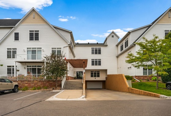 Front view of a multi-story white residential building on Old School Road, featuring an elevated entrance, stairs, ramp, and a small parking lot with vehicles in front of Unit 351 on the Mound. - Beise Real Estate