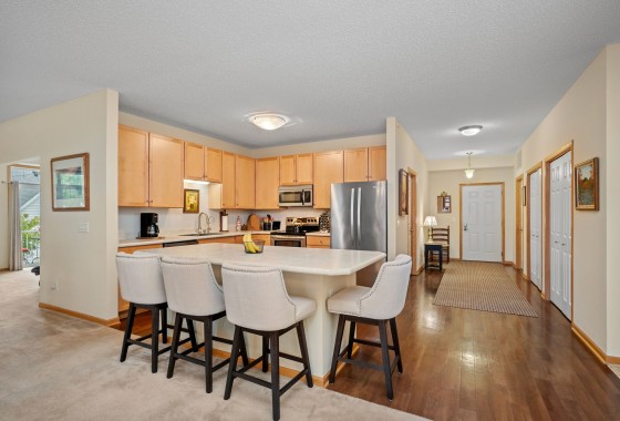 Modern kitchen in Mound with light wood cabinets, stainless steel appliances, a central island with four upholstered bar stools, and an adjacent open hallway with wood flooring for easy, maintenance free living in a walkable neighborhood. - Beise Real Estate