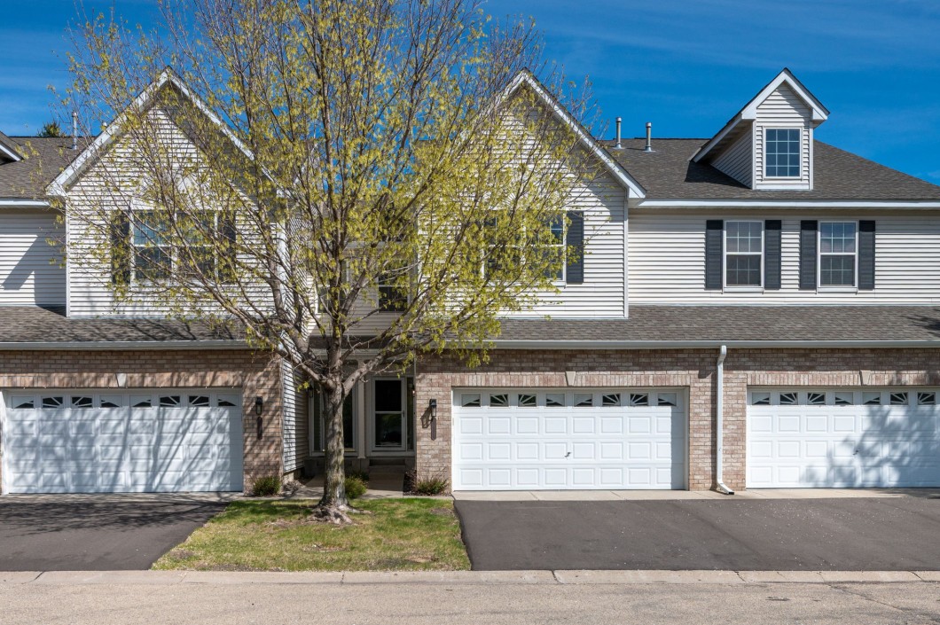 This Eden Prairie townhome features two stories, light siding with brick accents, three white garage doors, and a tree with sparse leaves in front. - Beise Real Estate
