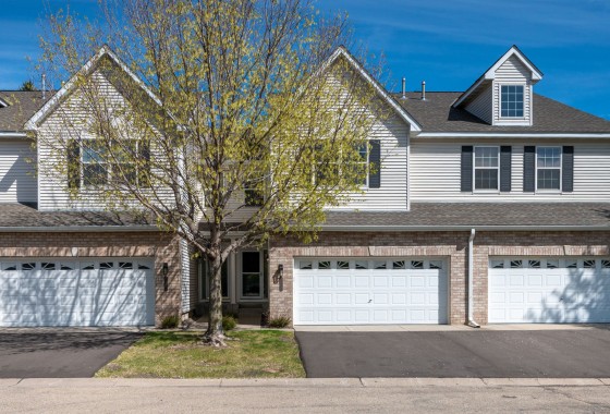 This Eden Prairie townhome features two stories, light siding with brick accents, three white garage doors, and a tree with sparse leaves in front. - Beise Real Estate