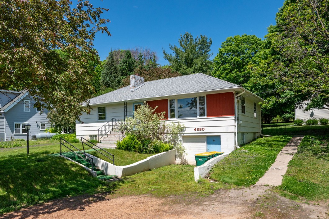 Charming single-story white house with a gray roof, red accents, and a front ramp at 4880 Grand View Avenue, Minnetrista—surrounded by trees and a lush green lawn on a sunny day. - Beise Real Estate