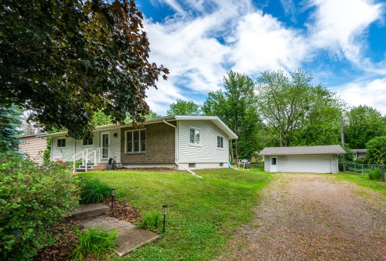 Charming single-story house on Rainbow Ave in Maple Plain with brick and siding exterior, surrounded by trees and grass. A gravel driveway leads to a detached garage beneath a partly cloudy sky. - Beise Real Estate