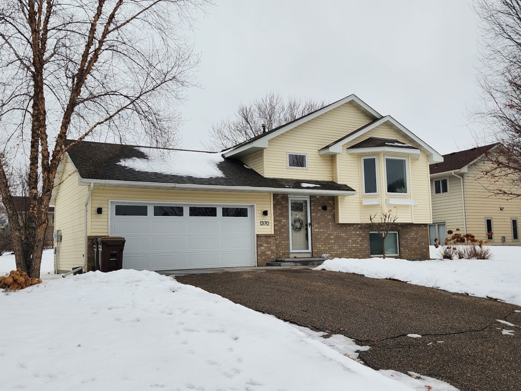 A split-level house at 1370 Fox Street in Delano features beige siding, brick accents, and a two-car garage. On this snowy, overcast day, the yard and driveway are blanketed in snow while leafless trees frame the scene. - Beise Real Estate