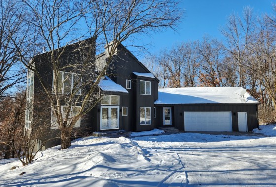 A modern two-story dark gray house with large windows and an attached white garage sits on Forest Ridge Drive in Lakeville, 22620, surrounded by leafless trees and snow under a clear blue sky. - Beise Real Estate