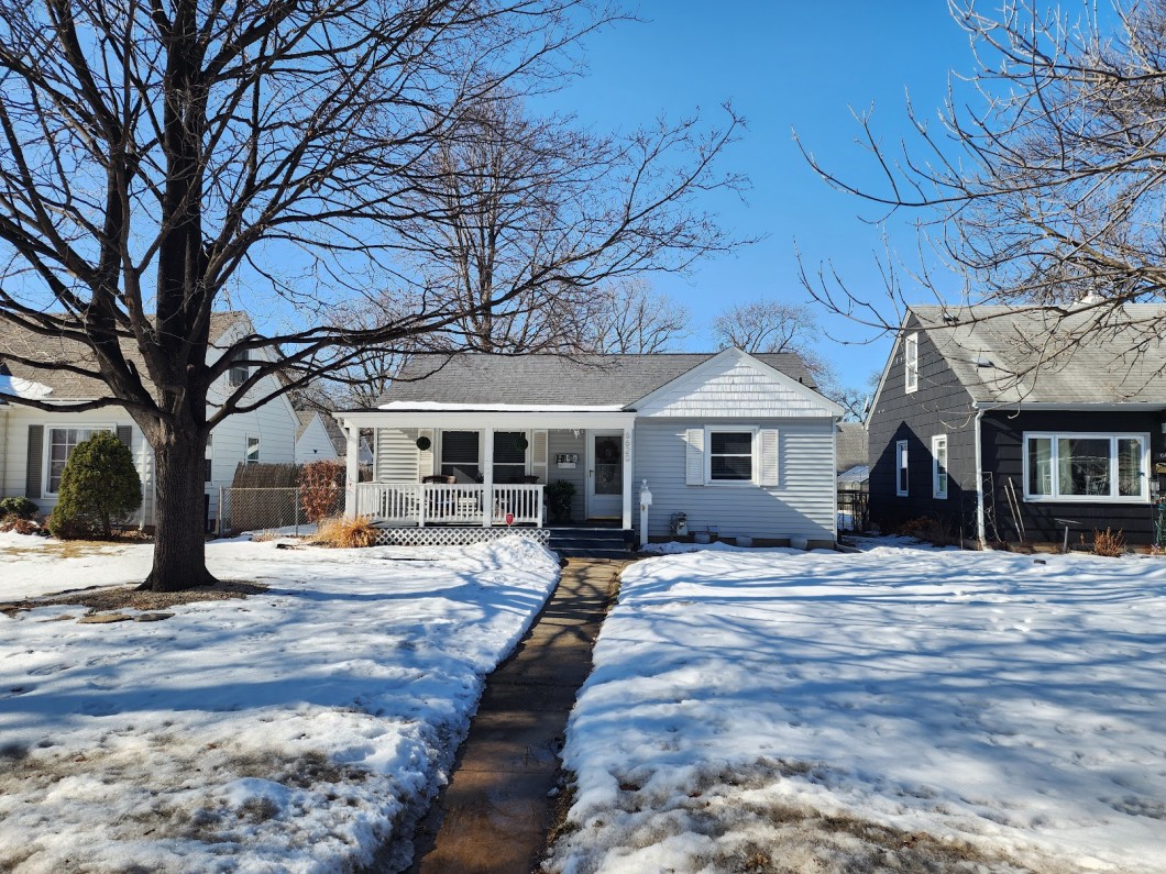 A small white house on 6620 Thomas Avenue in Richfield, with a front porch and steps, is surrounded by melting snow and bare trees on a sunny winter day. - Beise Real Estate