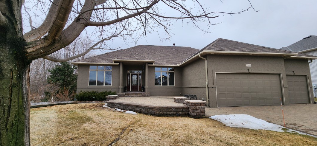 Single-story house in Maple Grove with gray stucco exterior, large windows, attached two-car garage, bare tree, and patches of snow on a brown lawn. - Beise Real Estate