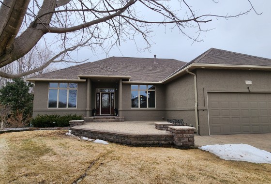 Single-story house in Maple Grove with gray stucco exterior, large windows, attached two-car garage, bare tree, and patches of snow on a brown lawn. - Beise Real Estate