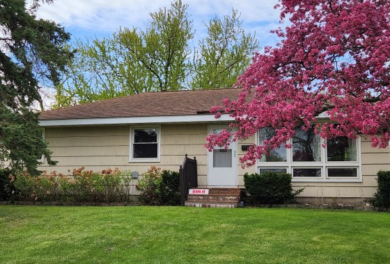 Charming single-story house at 902 7th Avenue E, Shakopee with beige siding, a brown roof, front lawn, blooming pink tree, and a "SOLD" sign on the steps. - Beise Real Estate